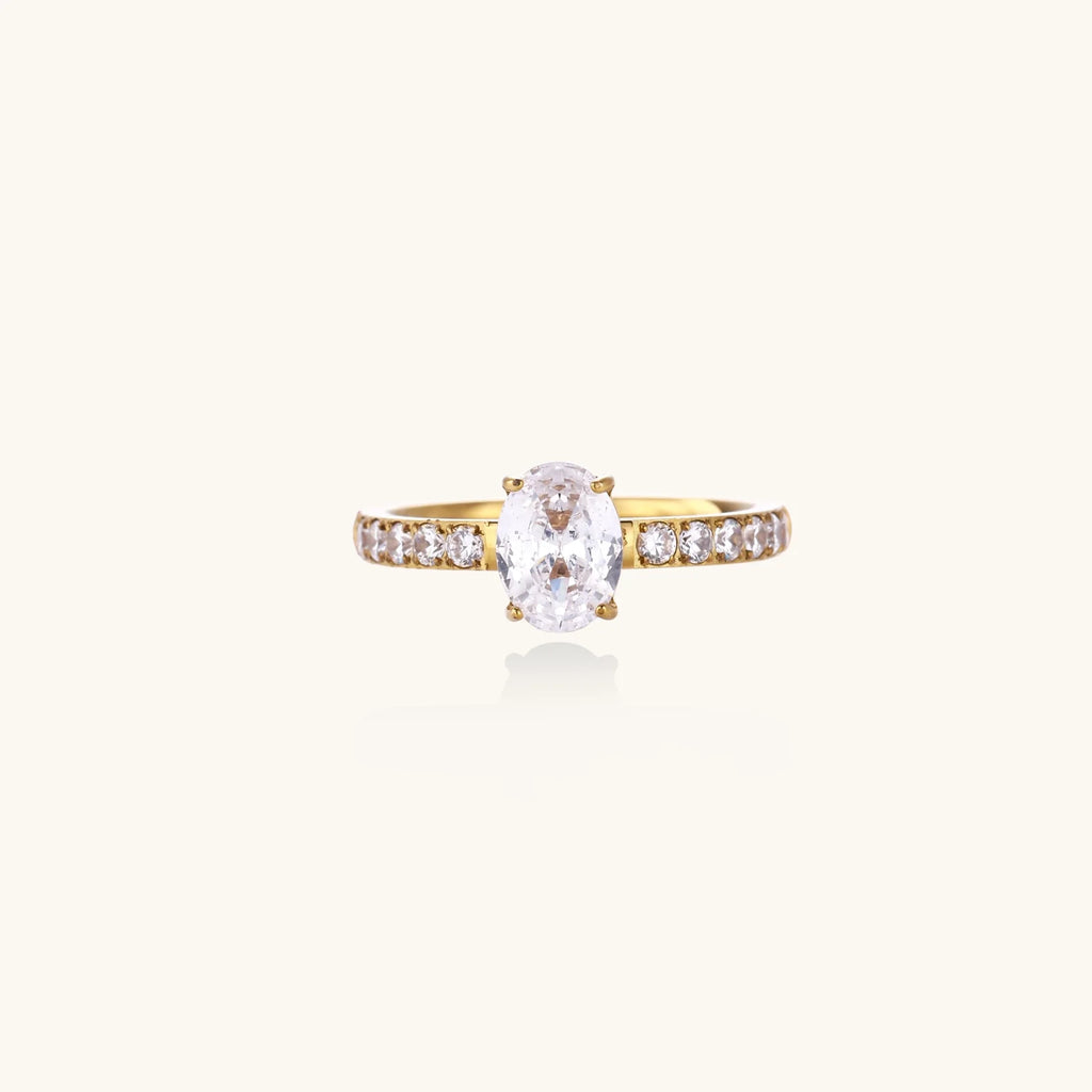 Gold-plated solitaire ring with oval zirconia center stone and pavé band on cream background
