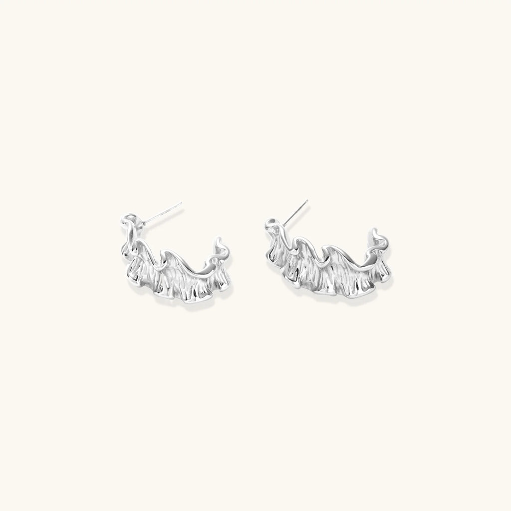 White gold plated sculptural hoop earrings with organic wavy edges and reflective metallic finish on cream background